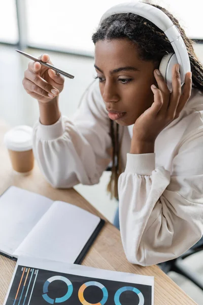 Vista de alto ángulo de la mujer de negocios afroamericana en auriculares sentados cerca de portátil y bebida para llevar en la oficina - foto de stock