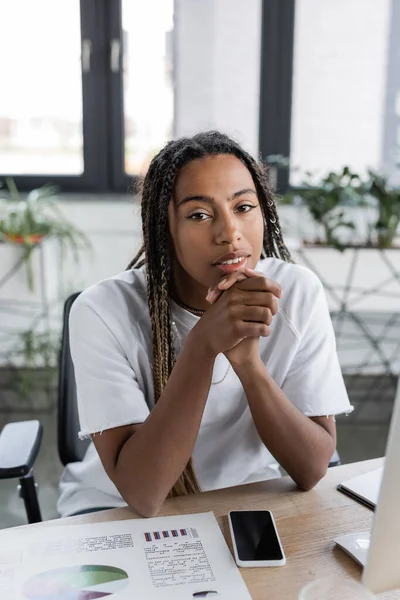 Femme d'affaires afro-américaine regardant la caméra près du smartphone et du papier avec des graphiques au bureau — Photo de stock