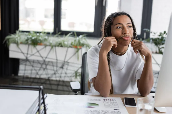 Femme d'affaires afro-américaine pensive touchant les cheveux près des appareils au bureau — Photo de stock