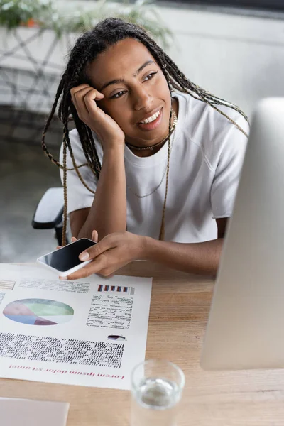 Mujer de negocios afroamericana sonriente sosteniendo un teléfono inteligente cerca del papel y la computadora en la oficina - foto de stock