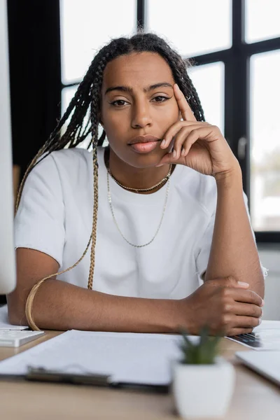 Retrato de una mujer de negocios afroamericana mirando la cámara cerca de un borroso portapapeles en la mesa en la oficina - foto de stock