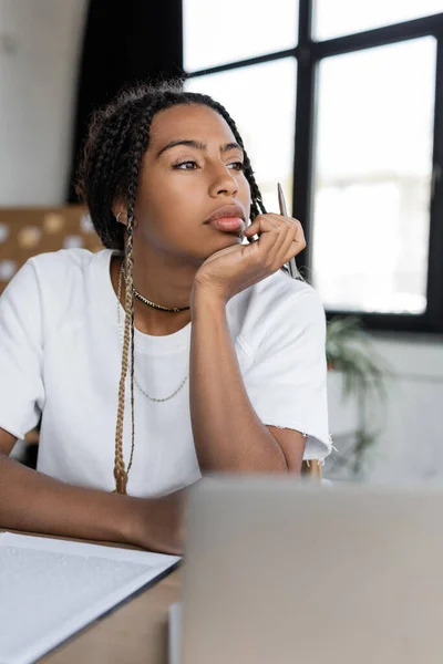 Empresária afro-americana em t-shirt segurando caneta perto da área de transferência e laptop borrado no escritório — Fotografia de Stock