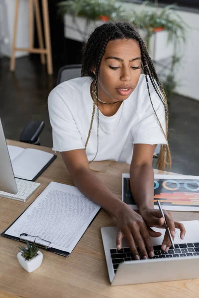 Vista de ángulo alto de la mujer de negocios afroamericana utilizando el ordenador portátil cerca de portapapeles y papel con gráfico en la oficina — Stock Photo