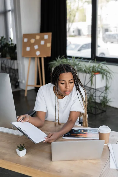 African american businesswoman in white t-shirt holding clipboard near devices and coffee in office — Stock Photo