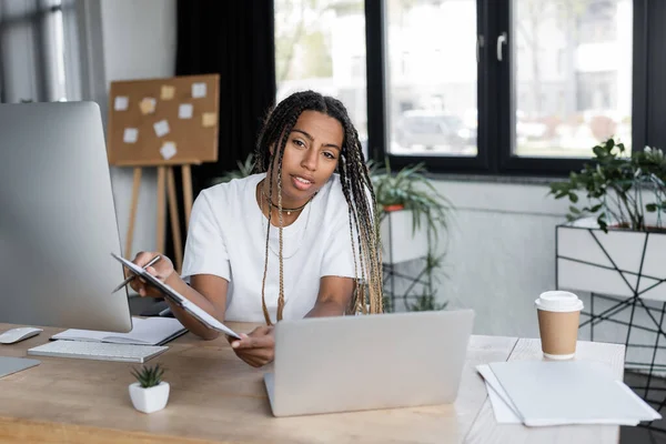 African american businesswoman holding clipboard and looking at camera near devices in office — Stock Photo