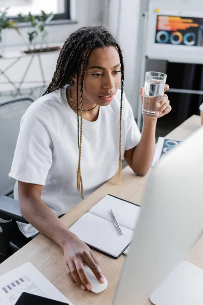 Femme d'affaires afro-américaine tenant un verre d'eau et utilisant un ordinateur dans un bureau moderne — Photo de stock