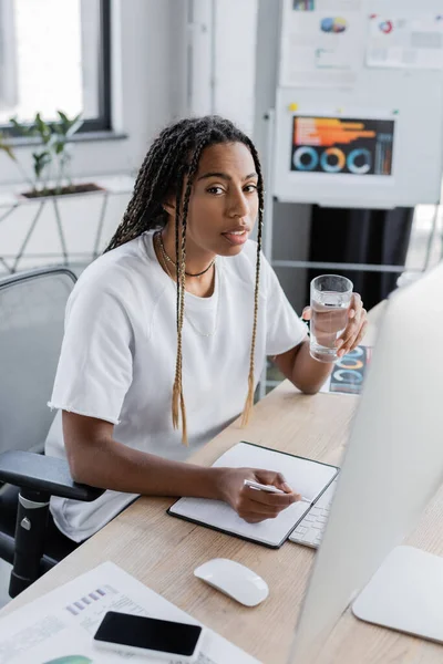 Femme d'affaires afro-américaine tenant un verre d'eau près du portable et de l'ordinateur au bureau — Photo de stock