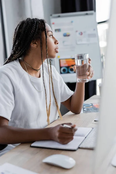 Vista laterale della donna d'affari afro-americana che tiene un bicchiere d'acqua vicino al notebook e al monitor del computer in ufficio — Foto stock