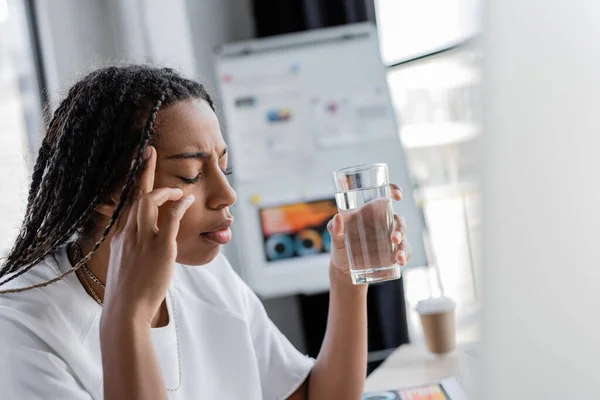 African american businesswoman holding glass of water while suffering from headache in office — Stock Photo