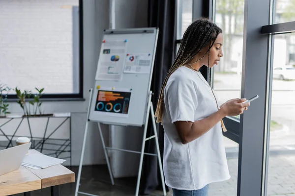 Vue latérale d'une femme d'affaires afro-américaine utilisant un smartphone près de la fenêtre dans un bureau moderne — Photo de stock