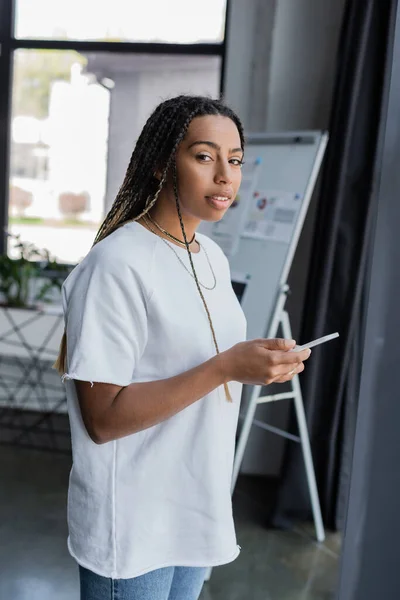 Mujer de negocios afroamericana en ropa casual sosteniendo el teléfono inteligente y mirando a la cámara en la oficina — Stock Photo