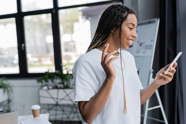 Positive african american businesswoman in t-shirt having video call on smartphone in office — Stock Photo