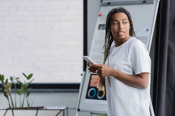 African american businesswoman in t-shirt holding smartphone near blurred flip chart in office — Stock Photo