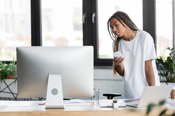 Afroamerikanische Geschäftsfrau im T-Shirt hält Smartphone neben Computer und Papiere im Büro — Stockfoto