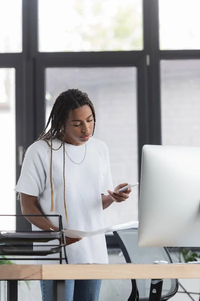 Femme d'affaires afro-américaine en vêtements décontractés tenant des papiers et utilisant un téléphone portable au bureau — Photo de stock