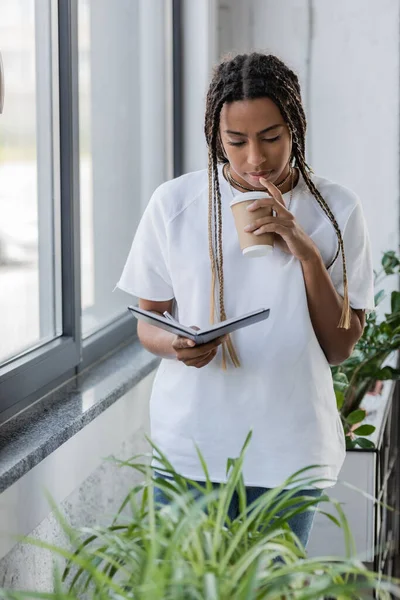 Femme d'affaires afro-américaine tenant du café pour aller et carnet près des plantes au bureau — Photo de stock