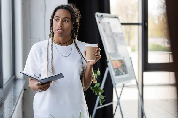 African american businesswoman holding notebook and takeaway drink in office — Stock Photo