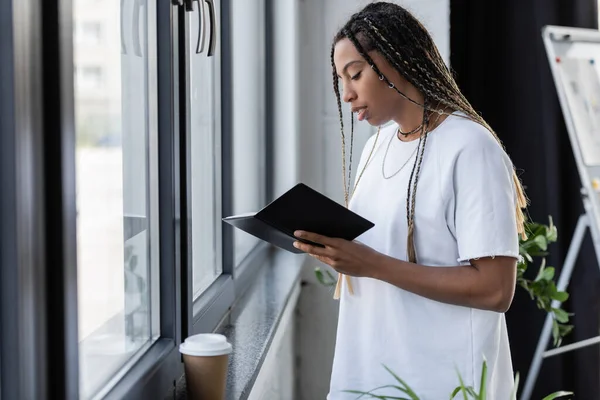 Joven mujer de negocios afroamericana sosteniendo cuaderno cerca de café para ir en alféizar de la ventana en la oficina - foto de stock