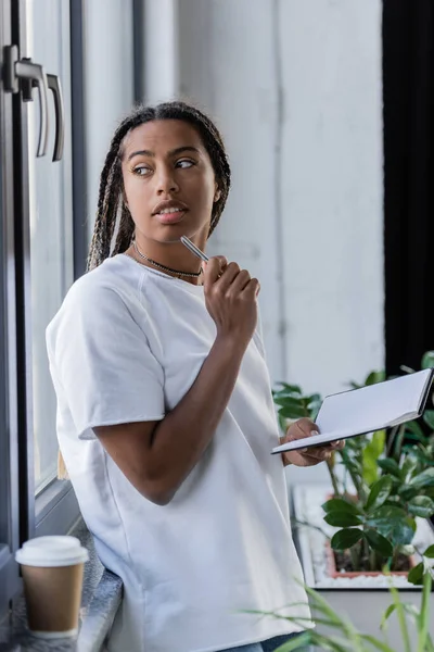 Mujer de negocios afroamericana en camiseta que sostiene la pluma y el cuaderno cerca del café en el alféizar de la ventana en la oficina - foto de stock