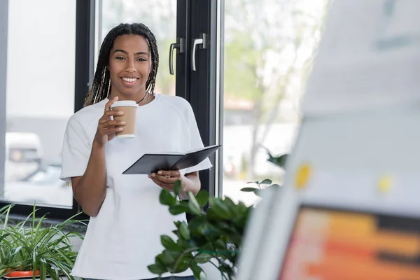 Positive afrikanisch-amerikanische Geschäftsfrau im T-Shirt mit Imbissgetränk und Notizbuch im Büro — Stockfoto