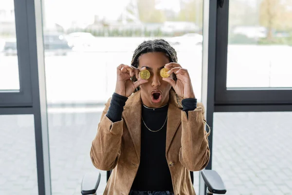 KYIV, UKRAINE - APRIL 27, 2022: Shocked african american businesswoman holding bitcoins near eyes in office — Stock Photo