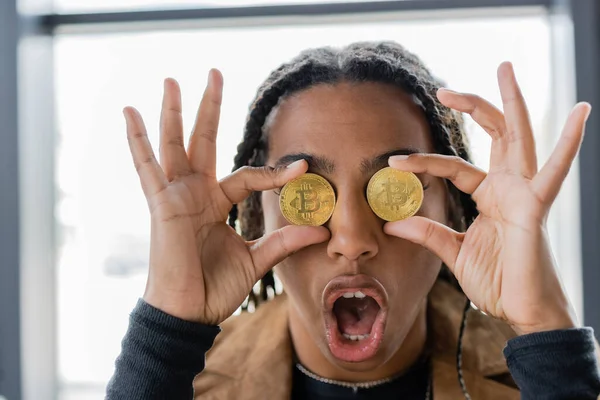 KYIV, UCRANIA - 27 de abril de 2022: Empresaria afroamericana conmocionada sosteniendo bitcoins cerca de los ojos en el cargo - foto de stock