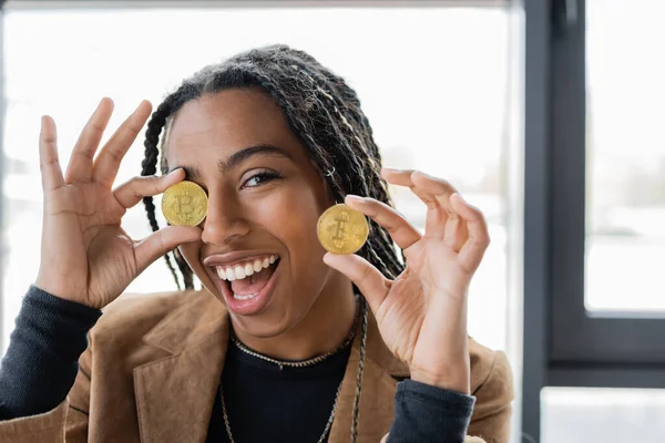 KYIV, UKRAINE - 27 AVRIL 2022 : Femme d'affaires afro-américaine joyeuse tenant Bitcoins en fonction — Photo de stock