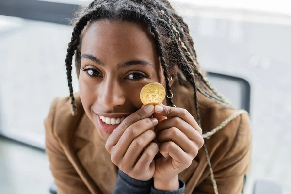 KYIV, UCRAINA - APRILE 27, 2022: Sorridente donna d'affari africana americana in possesso di bitcoin e guardando la fotocamera — Foto stock