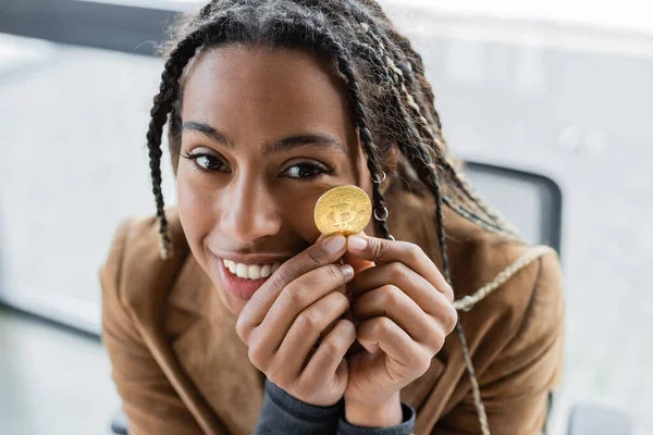 KYIV, UKRAINE - APRIL 27, 2022: Smiling african american businesswoman holding bitcoin and looking at camera — Stock Photo