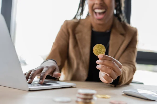 KYIV, UCRANIA - 27 de abril de 2022: Vista recortada de la borrosa mujer de negocios afroamericana sosteniendo bitcoin cerca de la computadora portátil en la oficina - foto de stock