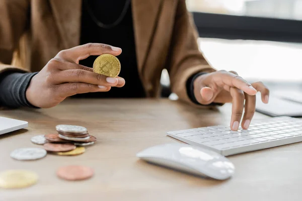 KYIV, UCRANIA - 27 DE ABRIL DE 2022: Vista recortada de la mujer de negocios afroamericana sosteniendo bitcoin y escribiendo en el teclado de la computadora - foto de stock