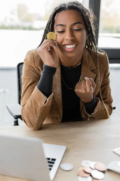 KYIV, UKRAINE - APRIL 27, 2022: Excited african american businesswoman holding bitcoin near laptop in office — Stock Photo