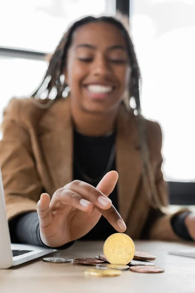 KIEW, UKRAINE - 27. April 2022: Verschwommene afrikanisch-amerikanische Geschäftsfrau berührt Bitcoin in der Nähe ihres Laptops im Büro — Stockfoto