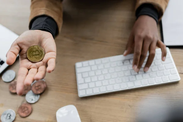 KYIV, UCRANIA - 27 DE ABRIL DE 2022: Vista recortada de la mujer de negocios afroamericana sosteniendo bitcoin y escribiendo en el teclado de la computadora - foto de stock