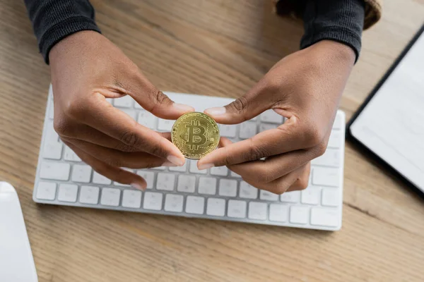 KYIV, UKRAINE - APRIL 27, 2022: Cropped view of african american businesswoman holding bitcoin near blurred computer keyboard in office — Stock Photo