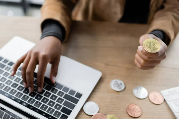 KYIV, UCRANIA - 27 DE ABRIL DE 2022: Vista recortada de la mujer de negocios afroamericana utilizando el ordenador portátil y la celebración de bitcoin en el cargo - foto de stock