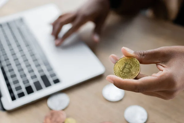 KYIV, UKRAINE - APRIL 27, 2022: Cropped view of african american businesswoman holding golden bitcoin and using laptop in office — Stock Photo