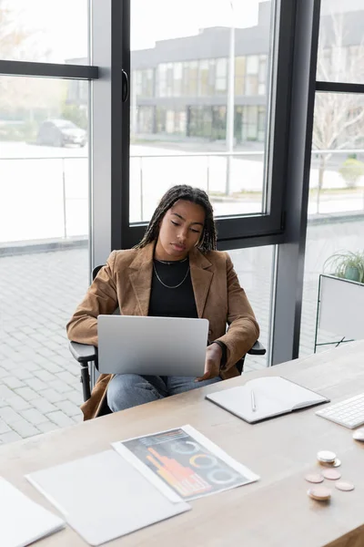 KYIV, UCRANIA - 27 DE ABRIL DE 2022: Empresaria afroamericana en chaqueta usando laptop cerca de papeles y notebook en oficina - foto de stock