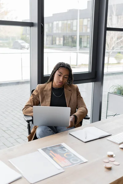 KYIV, UKRAINE - 27 AVRIL 2022 : Femme d'affaires afro-américaine vêtue d'un ordinateur portable près des papiers et d'un ordinateur portable au bureau — Photo de stock