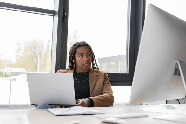 Mujer de negocios afroamericana con chaqueta que sostiene el ordenador portátil cerca del monitor de la computadora en la oficina - foto de stock