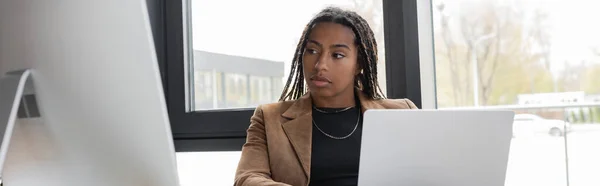 KYIV, UKRAINE - 27 AVRIL 2022 : Femme d'affaires afro-américaine regardant un écran d'ordinateur près d'un ordinateur portable au bureau, bannière — Photo de stock