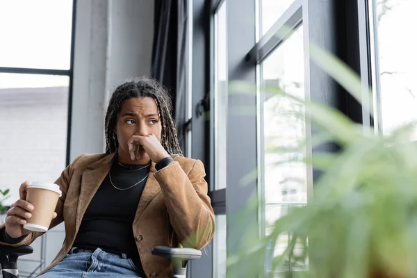 Preocupada afroamericana mujer de negocios en chaqueta celebración takeaway beber cerca ventana en oficina — Stock Photo