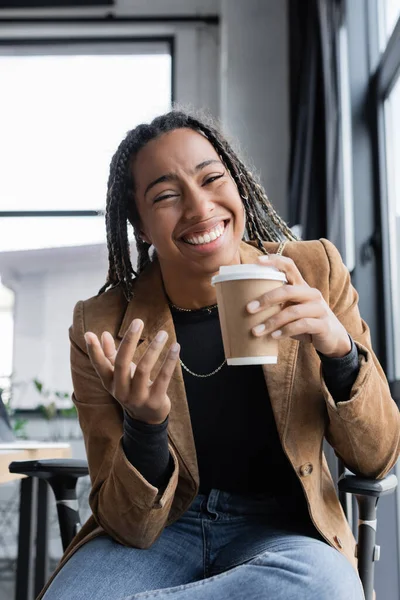 Positive african american businesswoman in jacket holding paper cup and looking at camera in office — Stock Photo