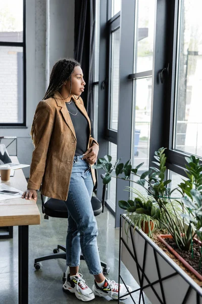 Vista lateral de la mujer de negocios afroamericana en chaqueta y jeans mirando a la ventana en la oficina - foto de stock