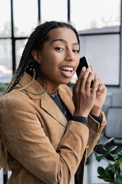 Positive afrikanisch-amerikanische Geschäftsfrau in Jacke hält Smartphone in der Hand und blickt in die Kamera im Büro — Stockfoto