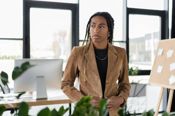 Mujer de negocios afroamericana con chaqueta mirando hacia otro lado en la oficina borrosa - foto de stock