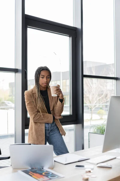 Donna d'affari afroamericana in possesso di caffè per avvicinarsi a dispositivi e notebook in ufficio — Foto stock