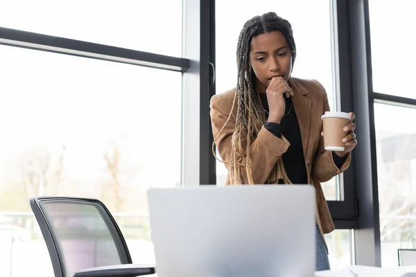Зосереджена афроамериканська комерсантка тримає каву, щоб піти і подивитися на ноутбук в офісі — стокове фото