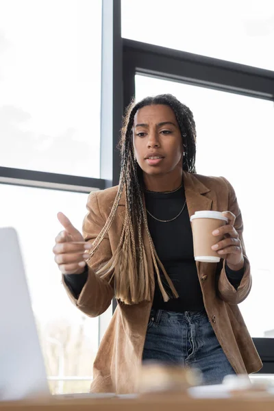 Femme d'affaires afro-américaine tenant un café pour aller et pointant du doigt à l'ordinateur portable dans le bureau — Photo de stock