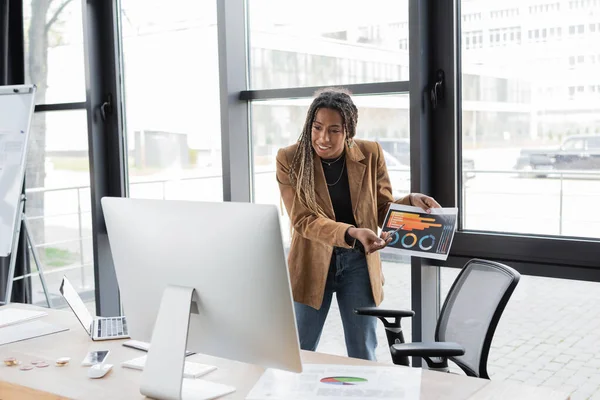 KYIV, UKRAINE - 27 AVRIL 2022 : Femme d'affaires afro-américaine tenant des graphiques lors d'un appel vidéo près de Bitcoins sur la table — Photo de stock